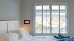 Ein ordentlich bezogenes Bett mit weißen Leinentüchern, einer Nachttischlampe und einem Fenster mit Jalousien.