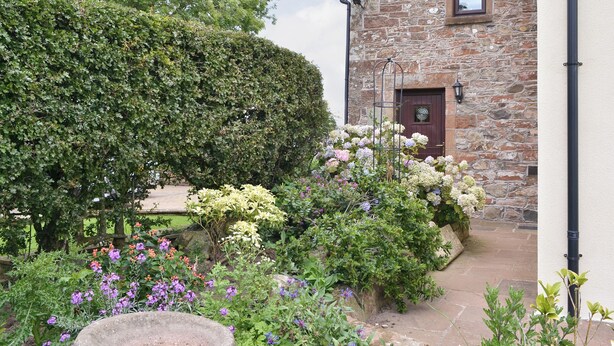 Ein Steinhaus mit Holztür und Fenster, umgeben von einem Garten mit verschiedenen Pflanzen und Blumen.