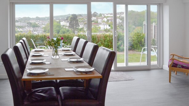 Uma sala de jantar com uma mesa grande posta para seis pessoas, janelas amplas com vista para uma área residencial e um banco de madeira no canto.