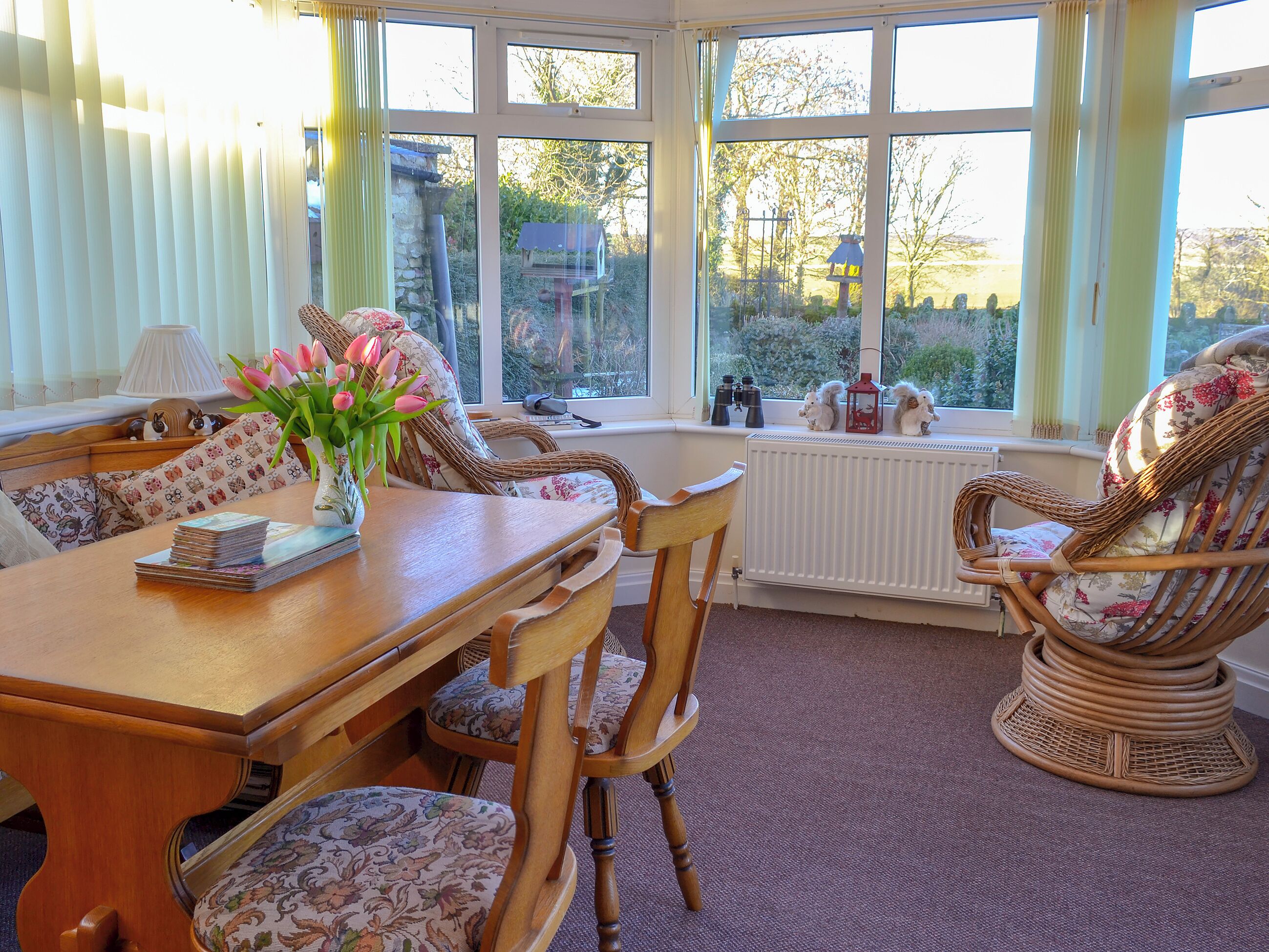 A sunlit room with a wooden table, wicker chairs, and a view of the garden through large windows.