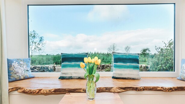 A wooden table with a vase of yellow tulips, two landscape paintings, and a bench in front of a window with a view of trees and sky.