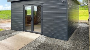 A small, dark-colored metal shed with a glass door, a concrete pathway leading to it, and a gravel area around.