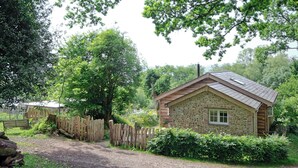 A stone cabin with a sloped roof, surrounded by a wooden fence and lush greenery.