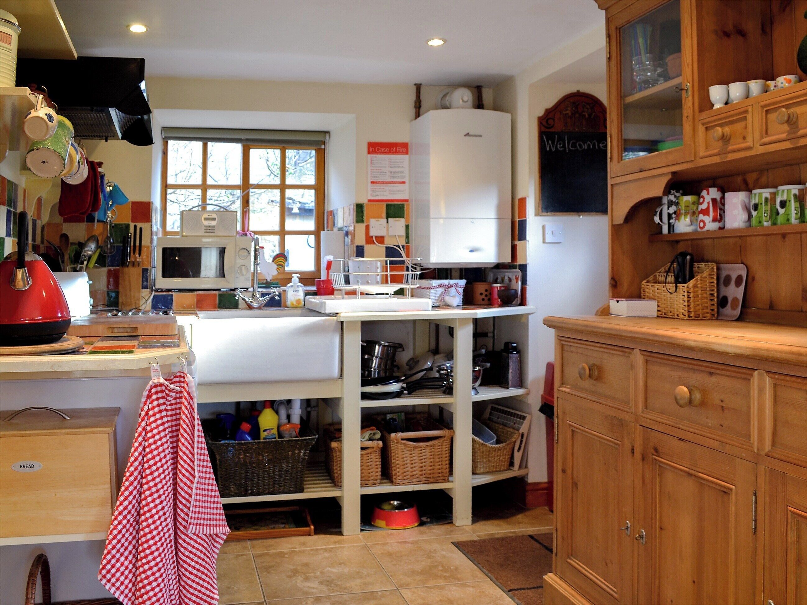 Eine Küche mit Mikrowelle, Herd und diversen Küchenutensilien. Es gibt ein Fenster mit Jalousien, einen Kühlschrank und Holzschränke.