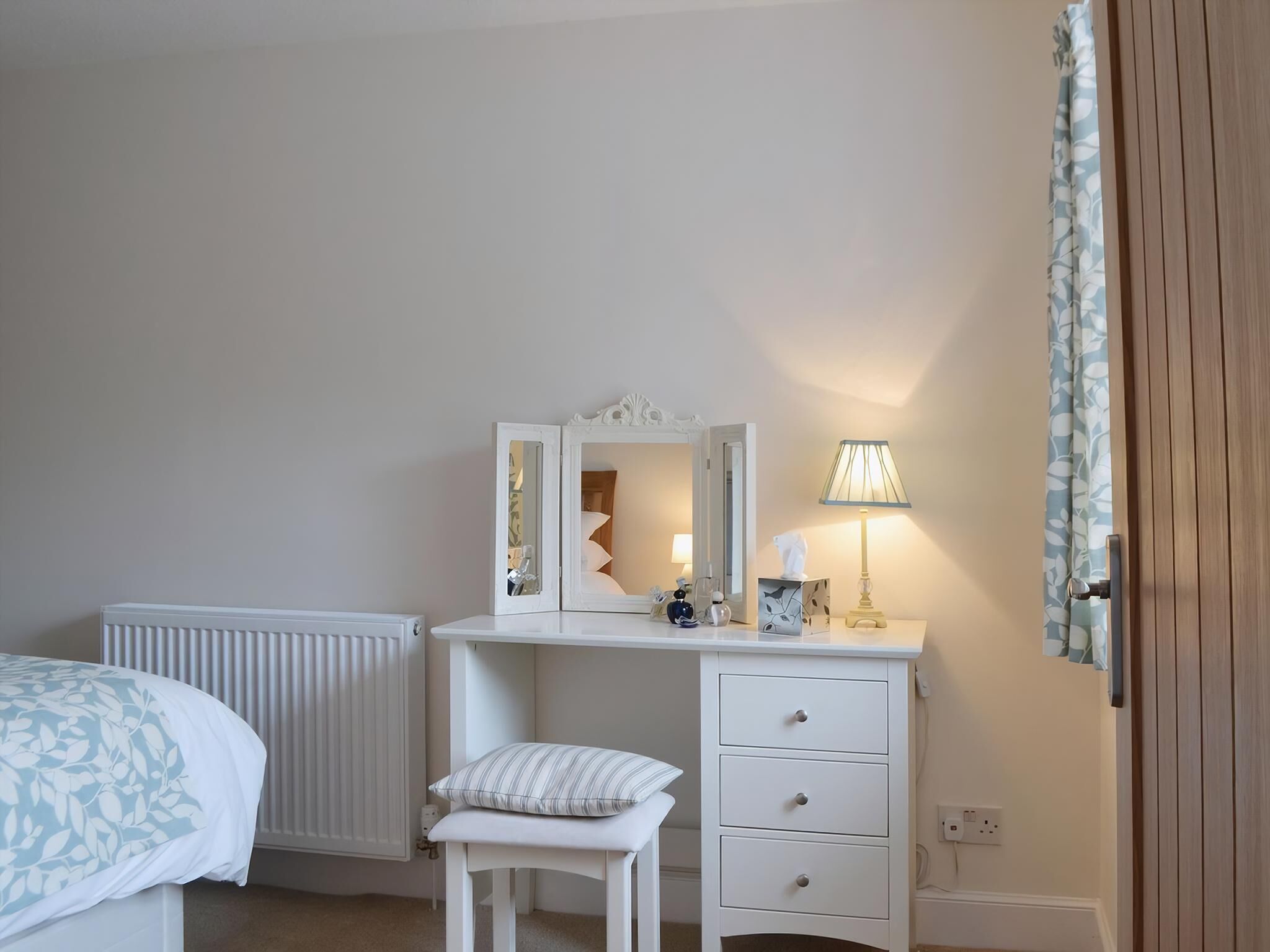 Une chambre à coucher avec un lit, une commode blanche à tiroirs, un miroir, une lampe et un radiateur.