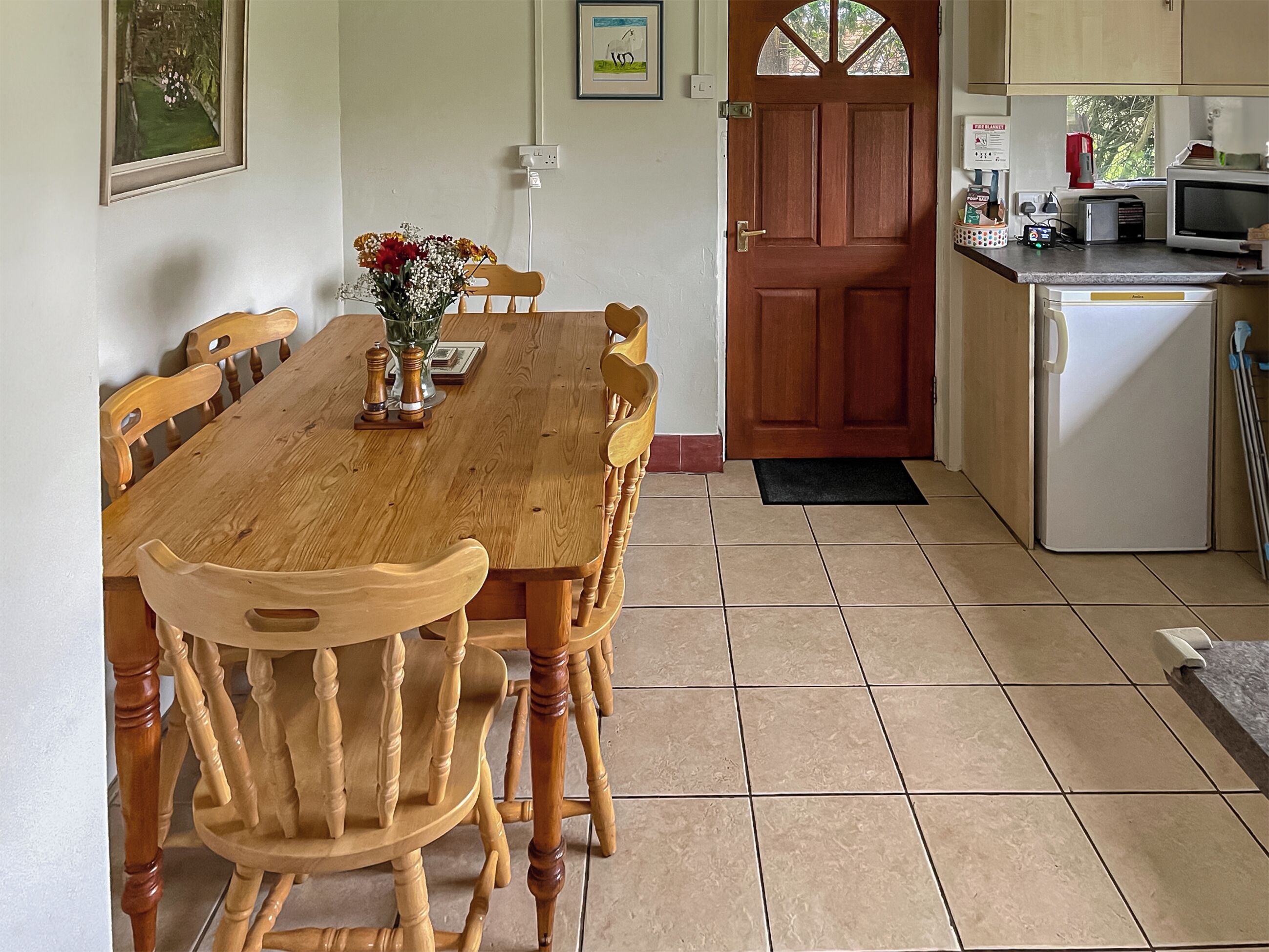 A dining area with a wooden table and chairs, a vase with flowers, and a kitchen with a microwave and toaster.