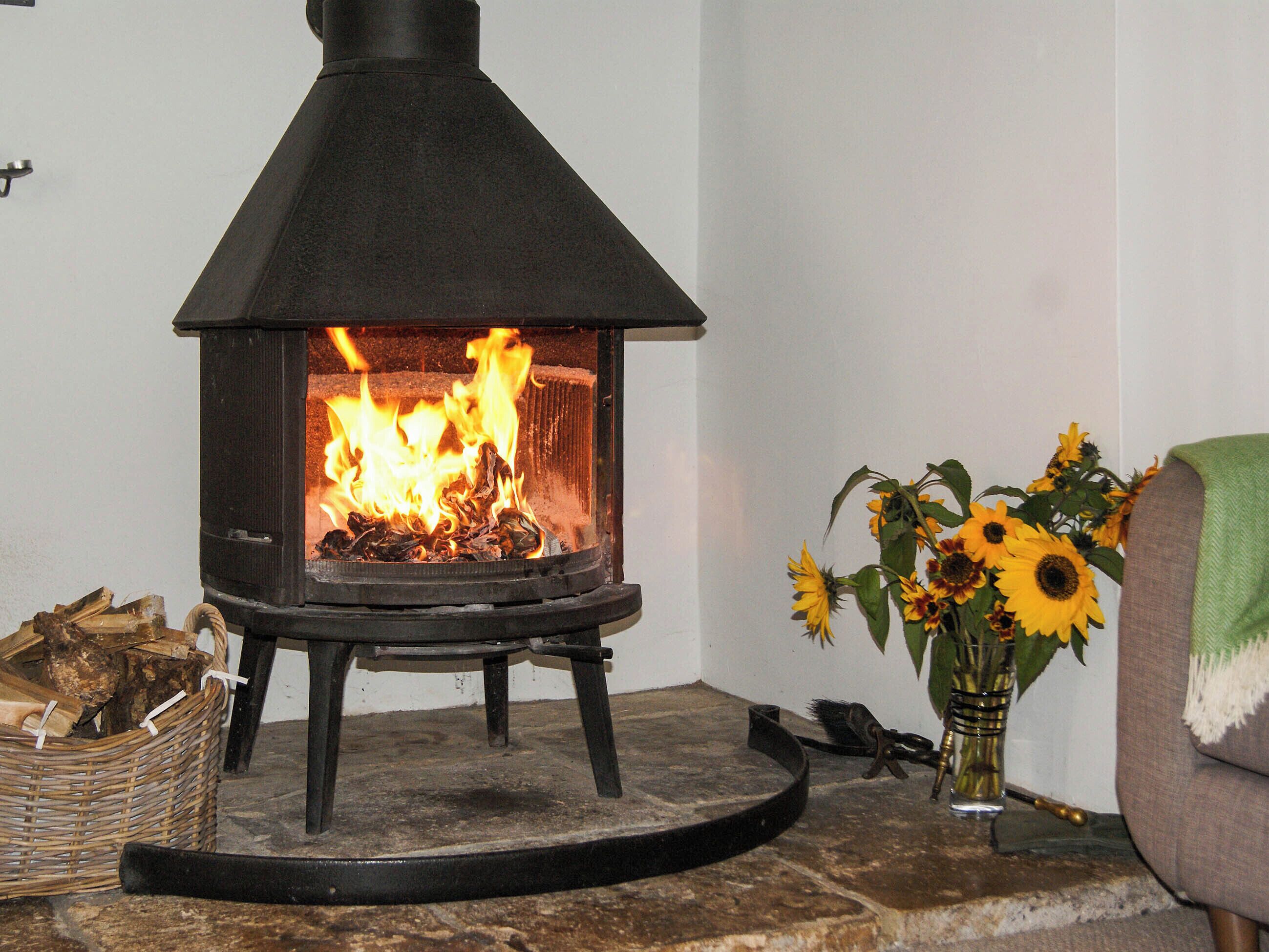 Un poêle à bois dans lequel le feu crépite, un panier en osier rempli de bois de chauffage et un vase de tournesols posé sur un sol en pierre.