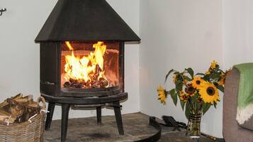 Un poêle à bois dans lequel le feu crépite, un panier en osier rempli de bois de chauffage et un vase de tournesols posé sur un sol en pierre.