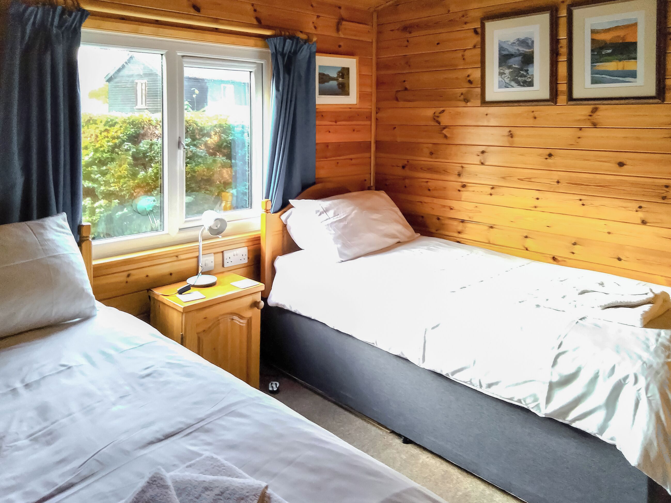 Et hyggeligt sommerhuskøkken med to senge, et natbord med en lampe og et vindue med gardiner.