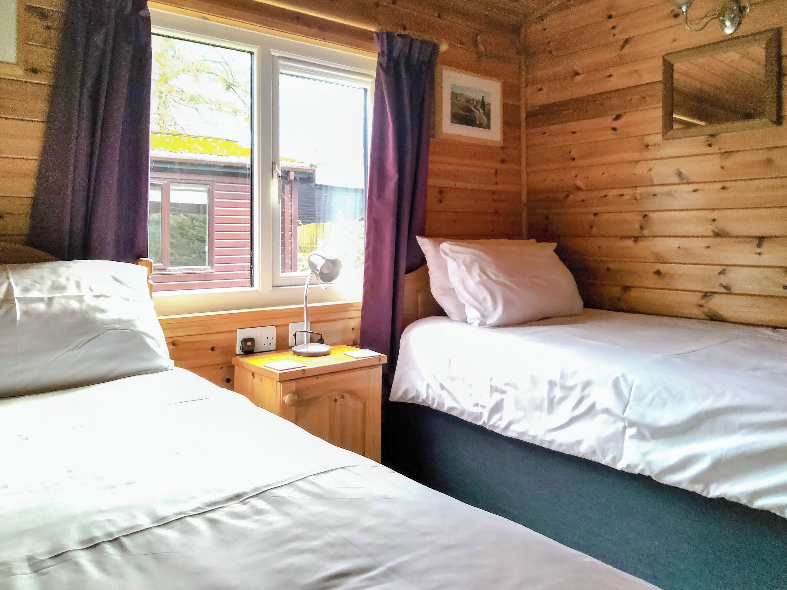 A wooden cabin room with two beds, a nightstand with a lamp, and a window with curtains.