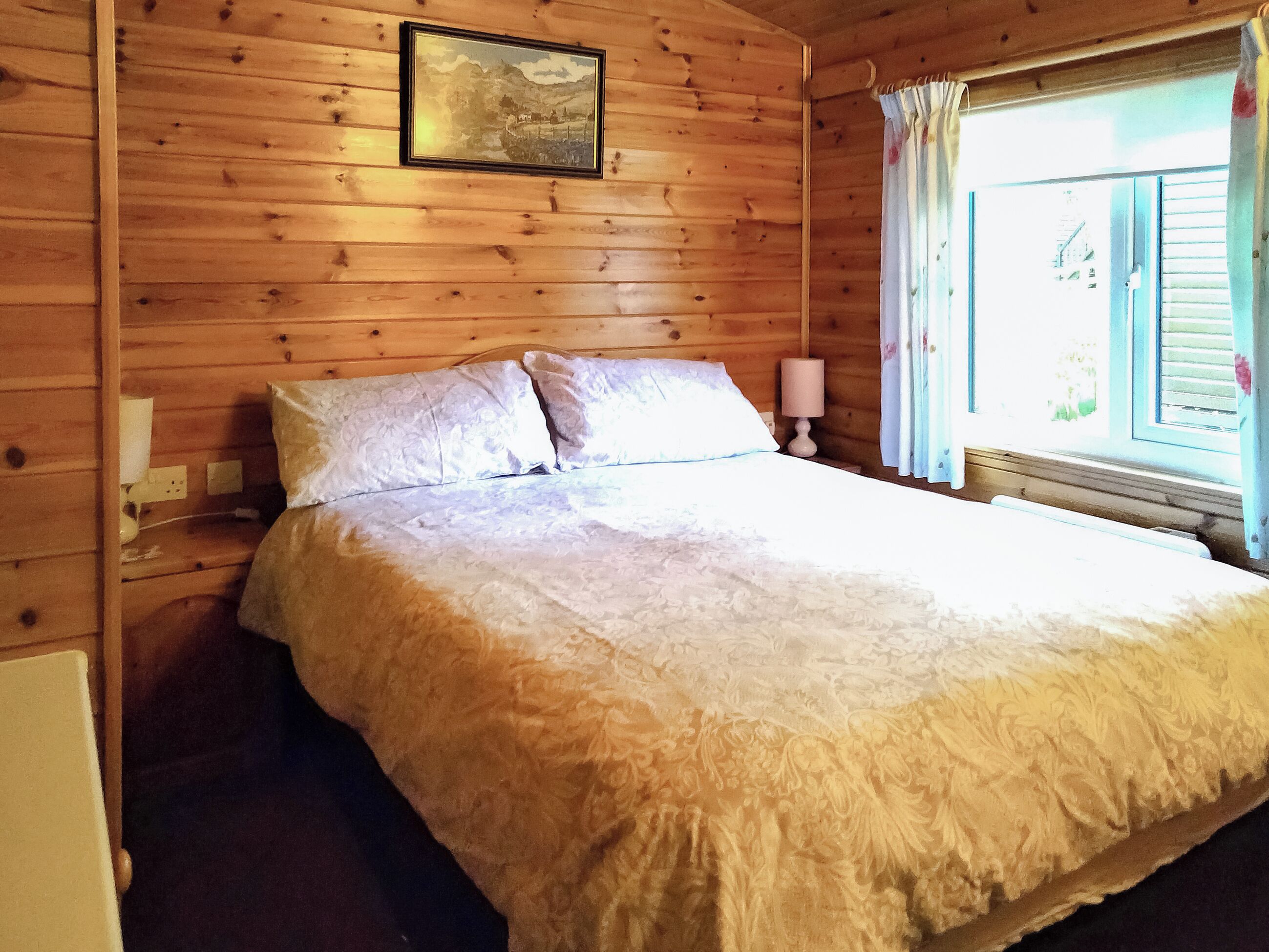 Una habitación de cabaña de madera con cama, mesita de noche, lámpara y ventana con cortinas.