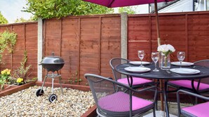 Une terrasse avec une clôture en bois, un barbecue et une table dressée pour les repas, agrémentée de coussins roses et d’un parasol violet.