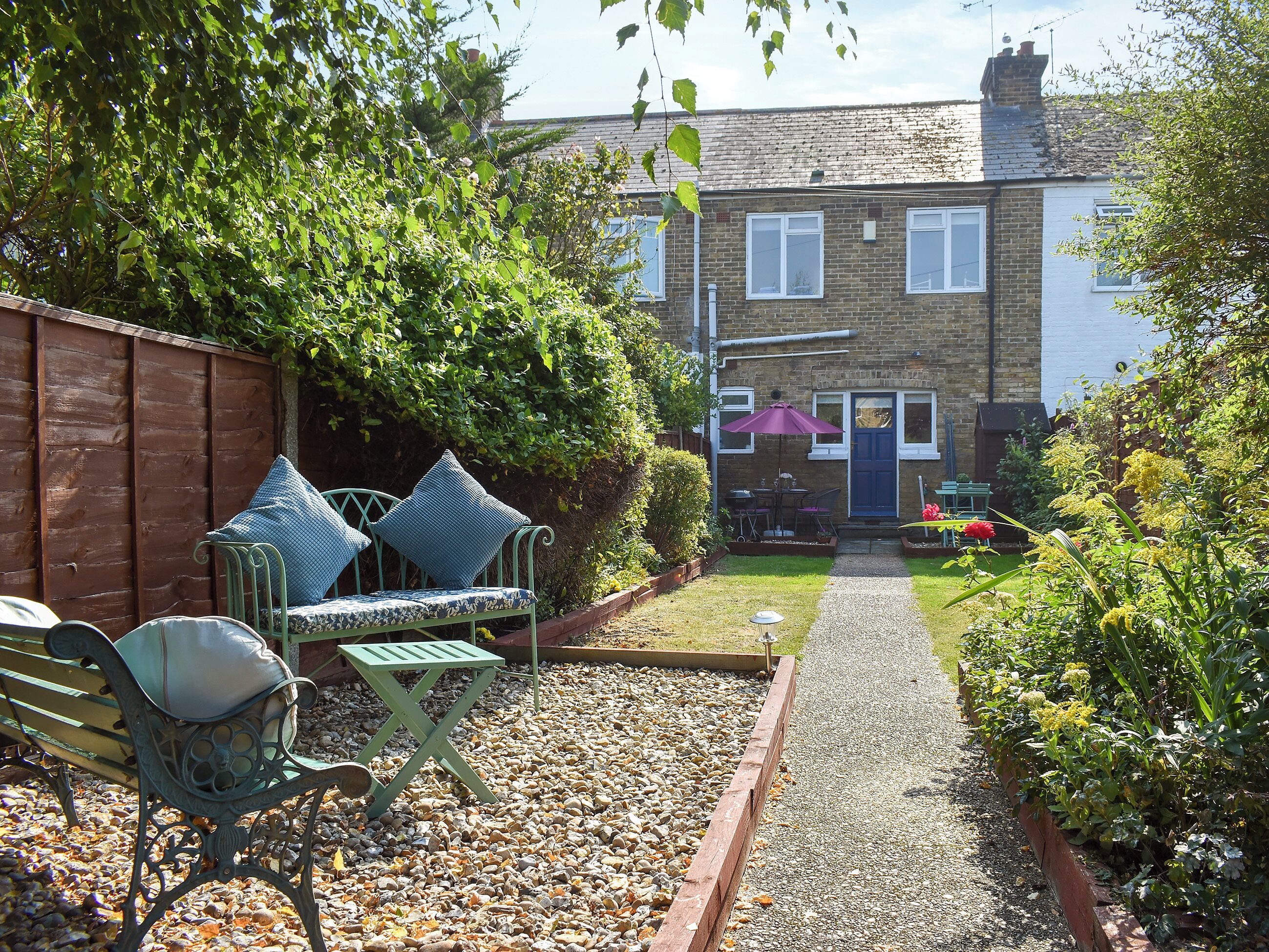 A garden with a gravel path leading to a house with a blue door. There is a bench with cushions and a table with chairs.