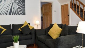 Un salon avec deux canapés gris foncé, dont l’un est agrémenté de coussins jaunes et noirs, et une table blanche sur laquelle est posée une plante. Une œuvre d’art à motifs zébrés orne le mur, et un escalier en bois est visible au fond.
