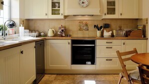 A kitchen with white cabinets, a dishwasher, an oven, and a dining area with a wooden table and chair. - 3 bedroom accommodation in Oldcroft, Lydney (Oldcroft, Lydney)