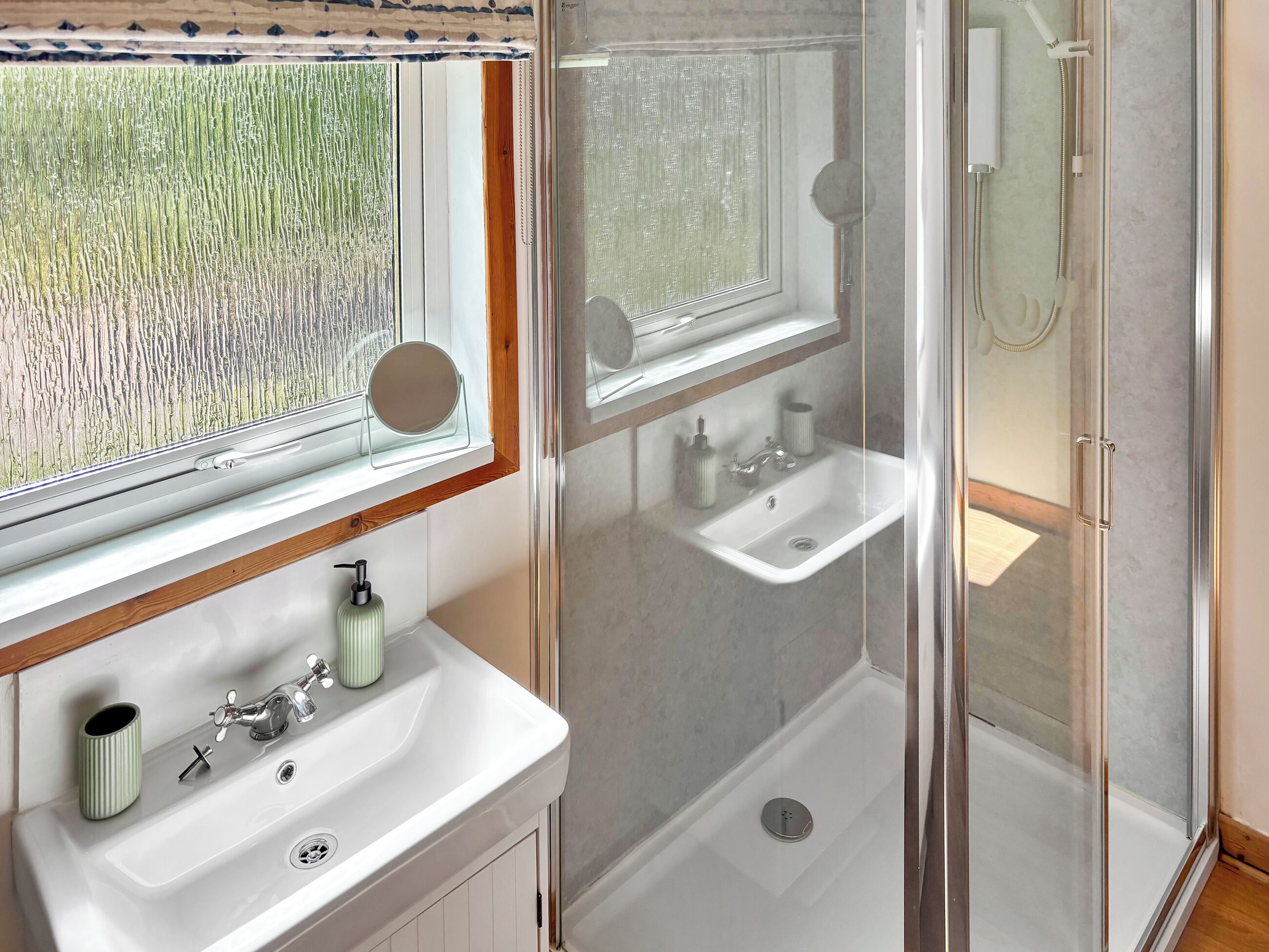 Een badkamer met een douchecabine van glas, een wastafel met spiegel erboven en een raam met uitzicht op groen.