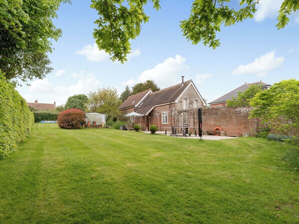 A brick house with a sloped roof, a well-maintained lawn, and a garden area with various plants and a shed. - 3 bedroom accommodation in Wareham (Wareham)