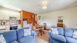 A compact living space with a kitchenette, two blue sofas with patterned cushions, a wooden dining table, and a staircase leading to an upper level. - Stable Cottage - 26032 (Wark, near Hexham)