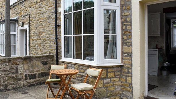 A stone building with a large bay window, a small outdoor seating area with two wooden chairs and a table, and a kitchen visible through an open doorway. - 2 bedroom accommodation in Skipton (Skipton)