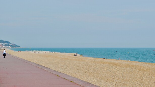 A coastal walkway with a pebbled beach, a clear sky, and a distant hill with buildings.