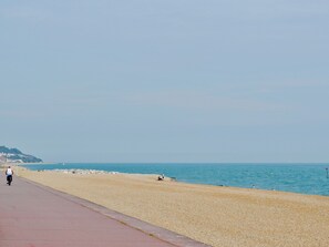 Un paseo marítimo con una playa de guijarros, cielo despejado y una colina distante con edificios.