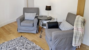 Una sala de estar con dos sillones grises, una mesita redonda con una lámpara, una alfombra y una pared decorada con cuadros.