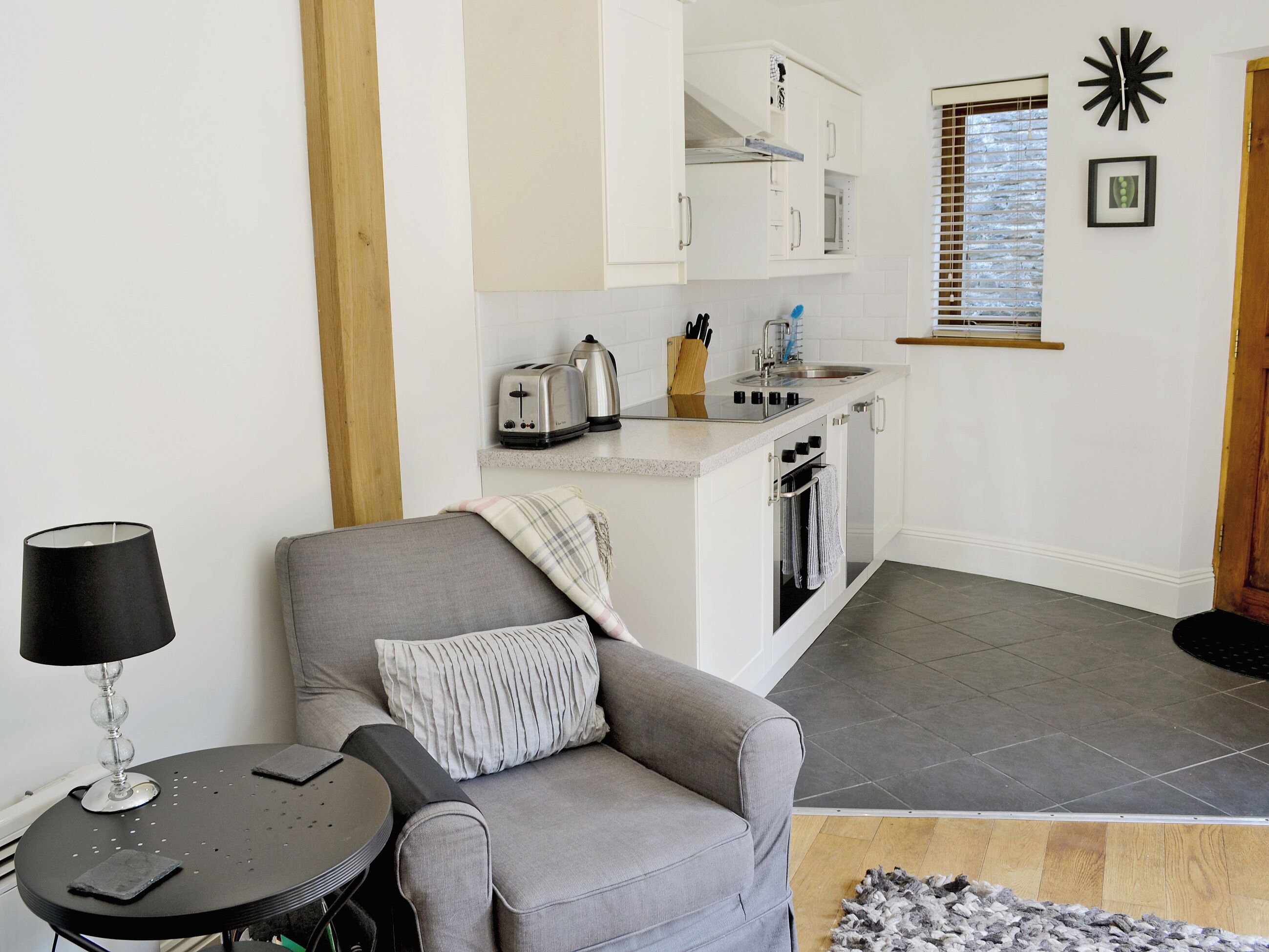 Un espacio habitable compacto con kitchenette, un sofá gris, una pequeña mesa redonda y una lámpara.