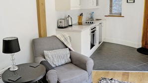 Un espacio habitable compacto con kitchenette, un sofá gris, una pequeña mesa redonda y una lámpara.