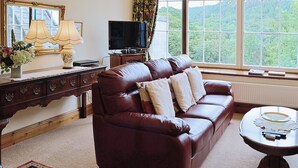 A living room with a leather sofa, a television, a table with remote controls, and a view of a forest through large windows.