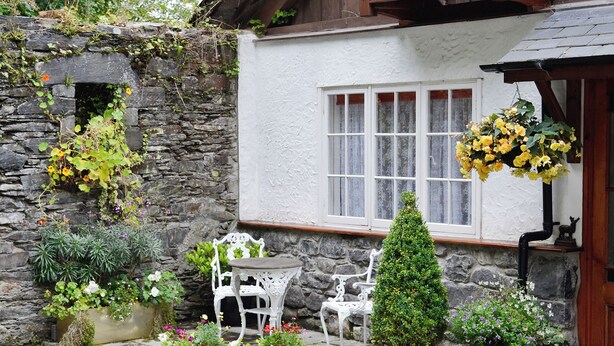 A stone wall with plants, a white chair, a small table, and a potted tree in front of a white building with a window.