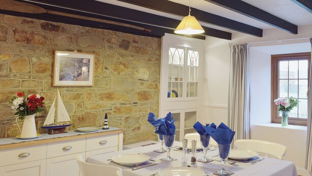 A dining area with a stone wall, a white cabinet, and a table set with plates, glasses, and blue napkins. - A beamed, character cottage situated just 100 yards from the River Teifi. (St Dogmaels near Cardigan)