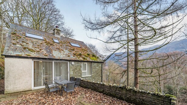 A stone house with a tiled roof, a patio with a table and chairs, and a view of mountains. - Super location within woodland and a SSSI. (Glenridding, near Penrith)