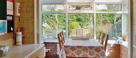A kitchen with a dining area, wooden cabinets, a table with a floral tablecloth, and a view of greenery outside.