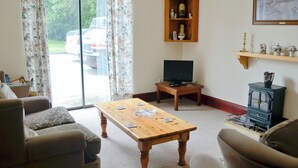 Een woonkamer met een houten salontafel, twee fauteuils, een televisie op een houten dressoir, een open haard en een raam met gordijnen.