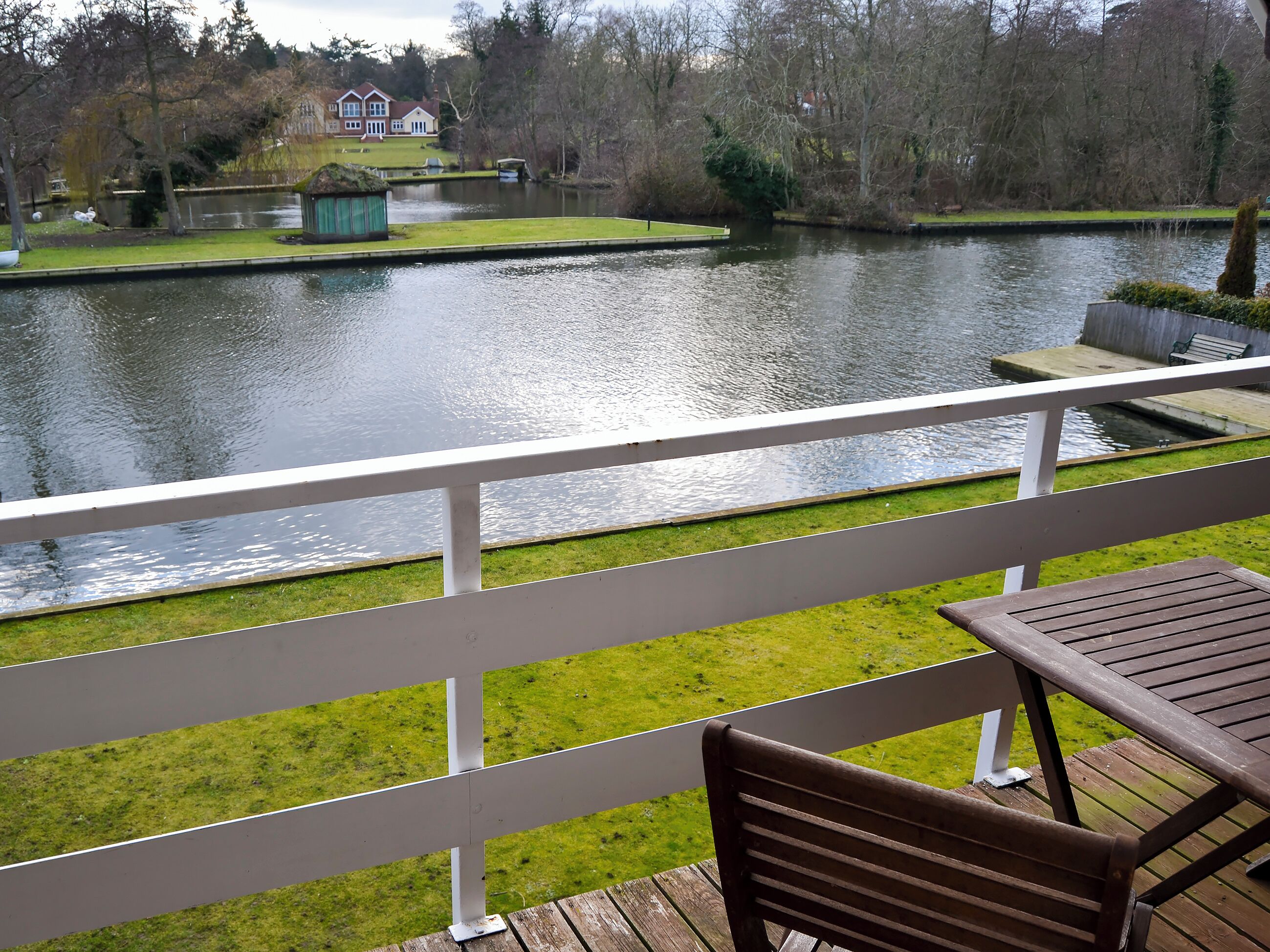 Un balcón con vistas a un río tranquilo, una zona verde y una casa a lo lejos.