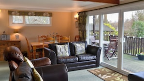 A cozy living room with a black leather sofa, a wooden dining table, and a large sliding glass door leading to an outdoor deck.