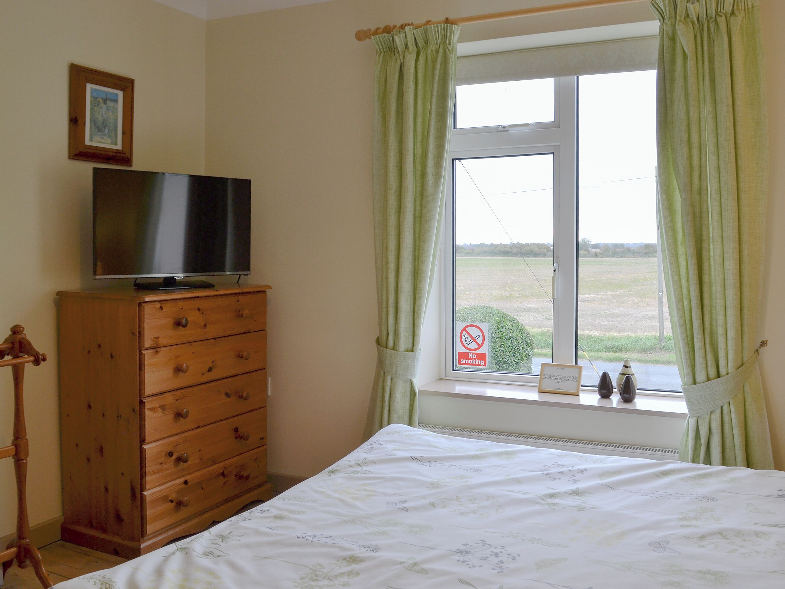 A bedroom with a bed, a wooden dresser, a television on top, and a window with a view of the outdoors.
