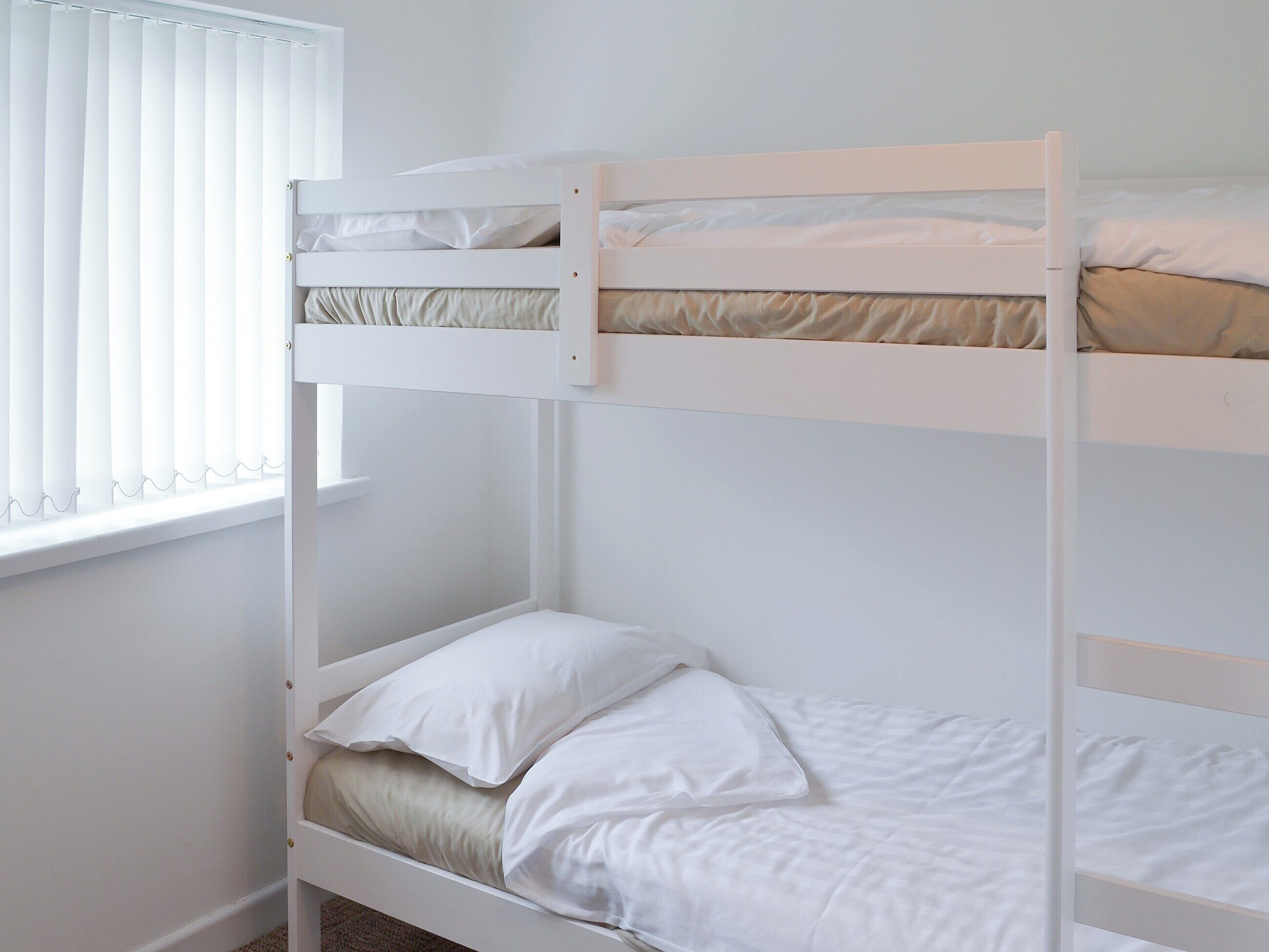 A bunk bed with white bedding in a room with white walls and a window with blinds.