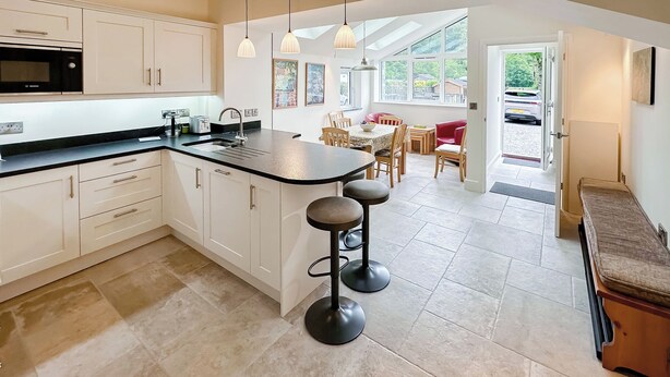 Een moderne keuken met witte kasten, een zwart aanrecht en een centraal eiland met krukken. Er is een zithoek met een rode stoel en een tafel, en een groot raam met uitzicht op buiten.