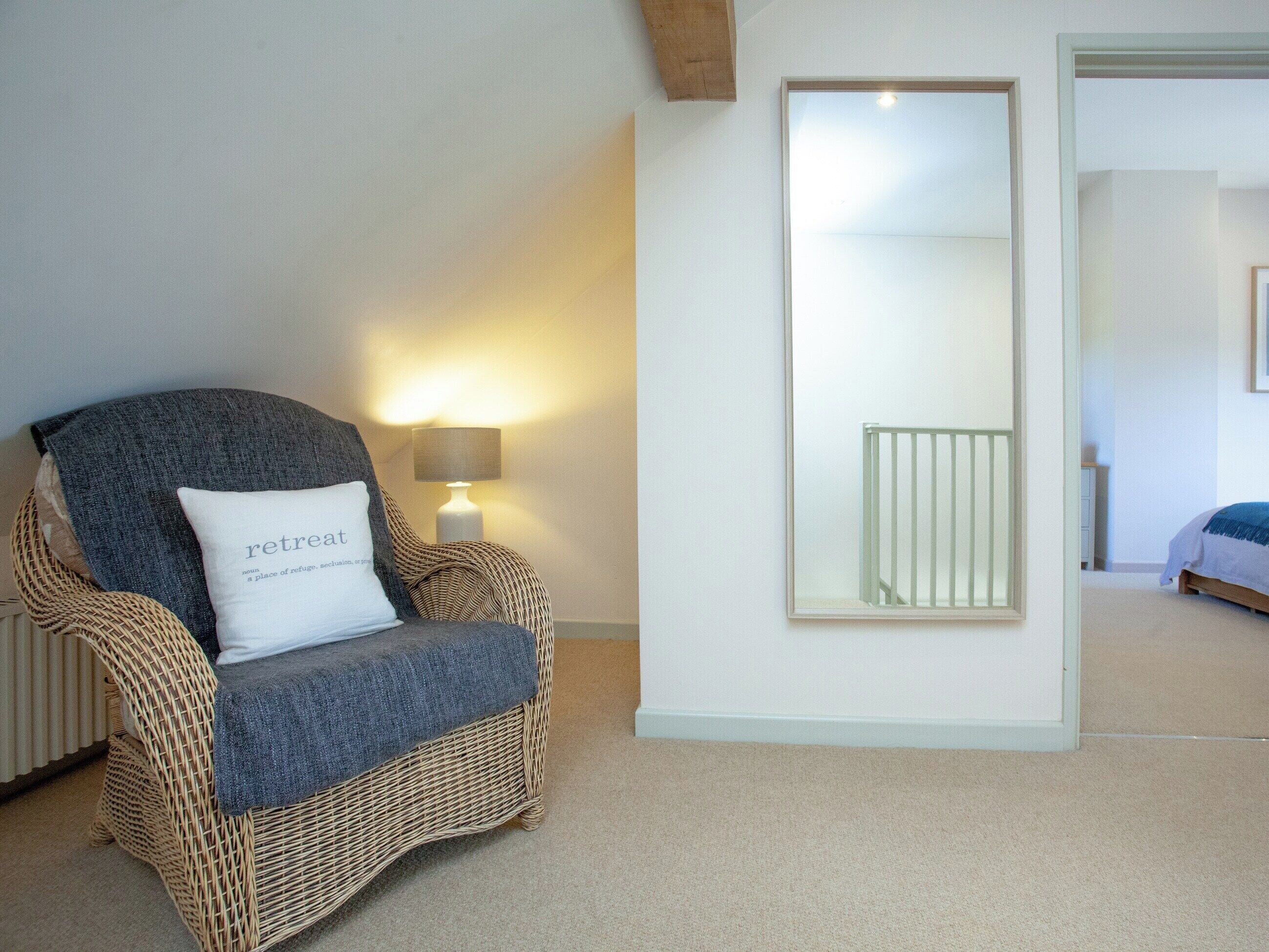 A wicker chair with a cushion labeled "retreat" and a lamp in a room with a large mirror and a bed in the background.