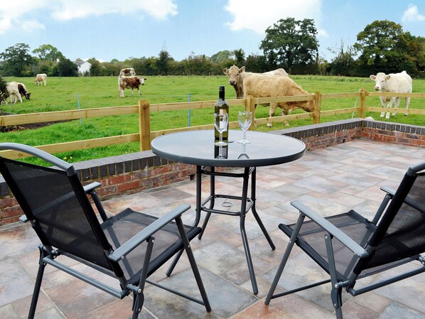 An outdoor patio with a round table and two chairs, a wooden fence, and cows in the background. - 1 bedroom accommodation in Weetwood, near Kelsall (Weetwood, near Kelsall)