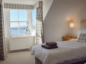 A bedroom with a bed, a nightstand, a lamp, a window with curtains, and a view of the sea. - Harbourside Apartment (Anstruther)