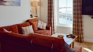 A living room with a sofa, a table with fruit, a lamp, and a view of the sea through a window. - Harbourside Apartment (Anstruther)
