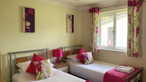 Una habitación con dos camas individuales, cada una con una colcha rosa y almohadas a juego. Hay dos lámparas de pared sobre mesitas de noche de madera, y una ventana con cortinas estampadas de flores.