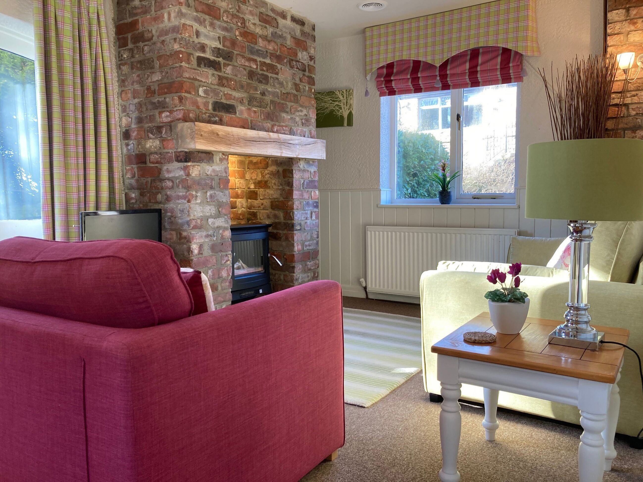 Een gezellige woonkamer met een haard van baksteen, een rode bank, een crèmekleurige fauteuil en een tafeltje met een lamp en bloemen.