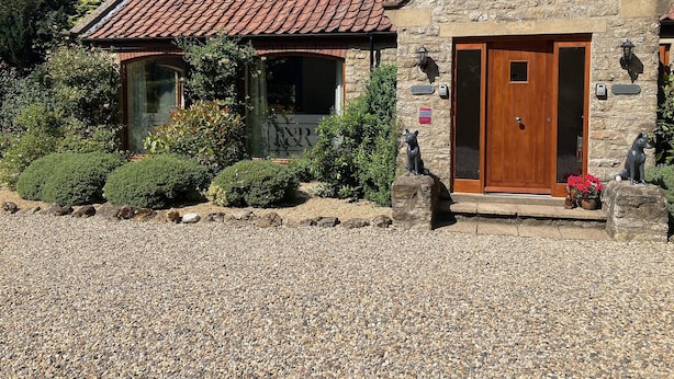 Un bâtiment en pierre avec un toit en tuiles, une porte en bois et un petit jardin agrémenté de galets et d’arbustes.