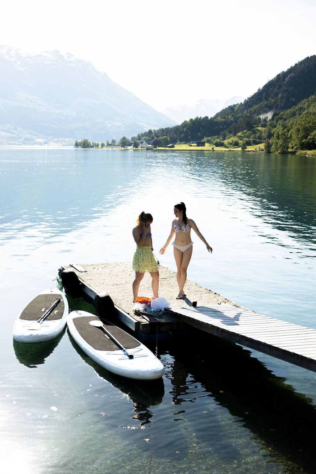 Two people on a dock with paddleboards, a calm lake, and mountains in the background.