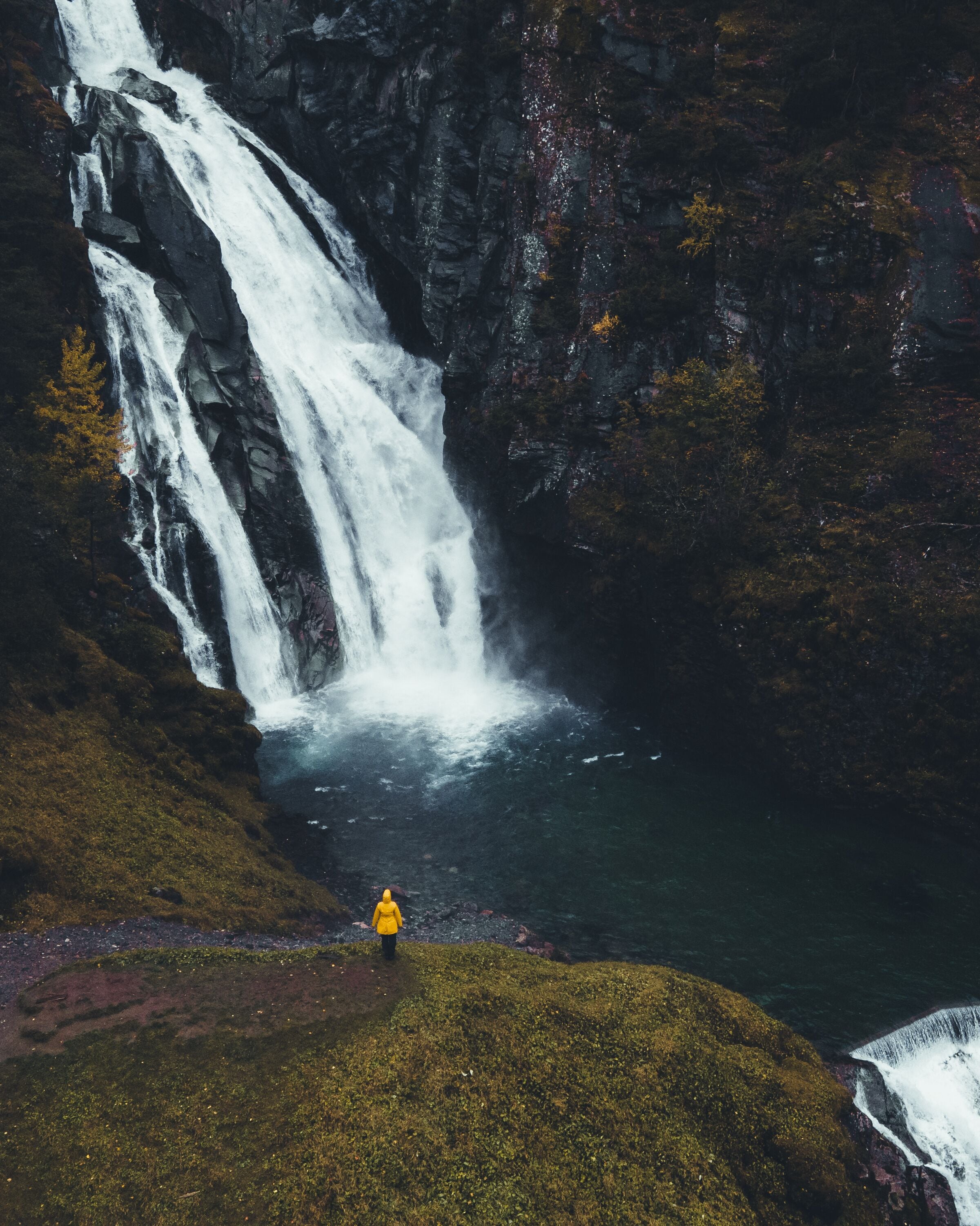 A waterfall cascading down a rocky cliff into a body of water, with a person in a yellow jacket standing on a grassy area at the base.