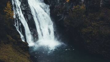 A waterfall cascading down a rocky cliff into a body of water, with a person in a yellow jacket standing on a grassy area at the base.