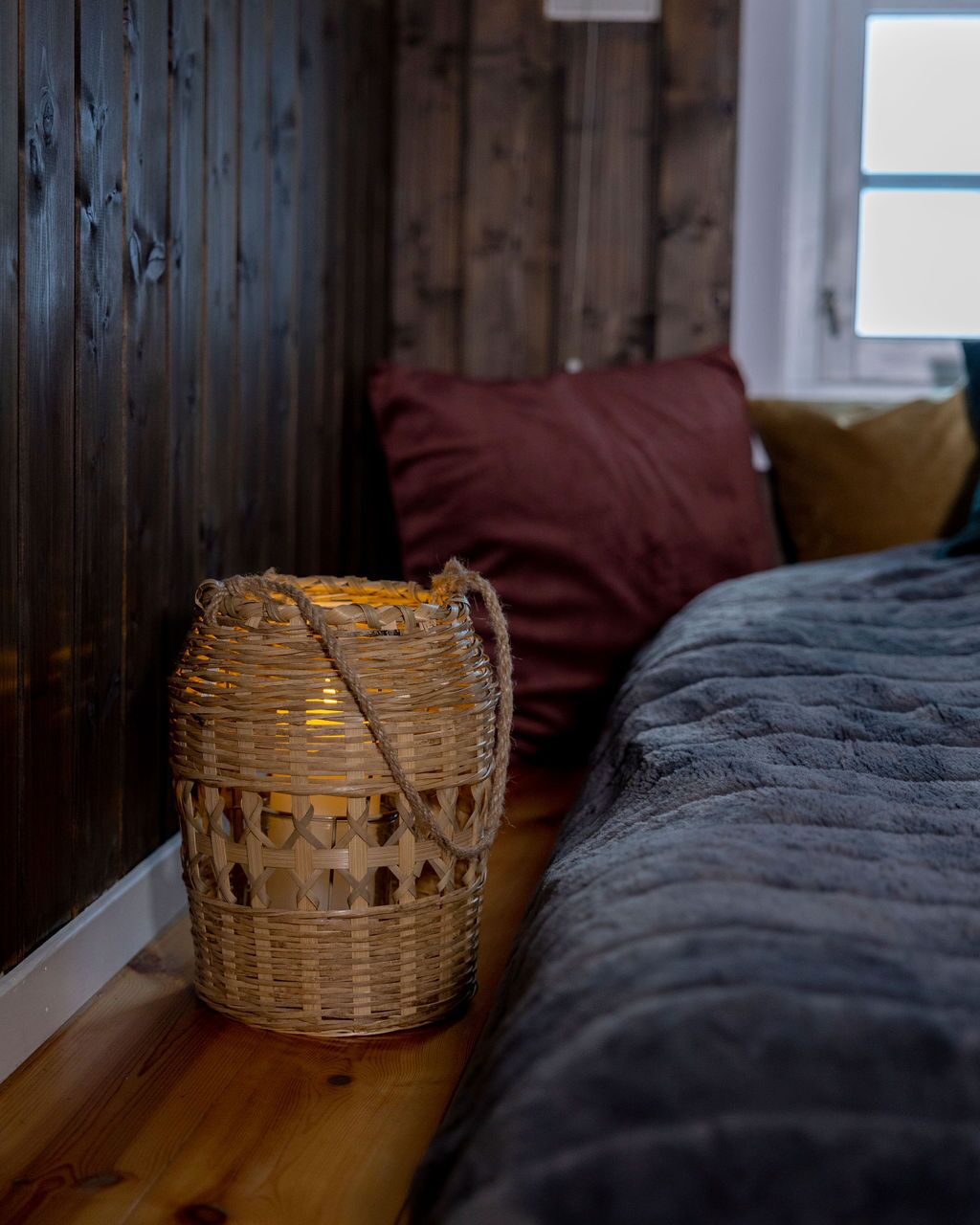 A wicker basket on a wooden floor next to a bed with a dark gray blanket.